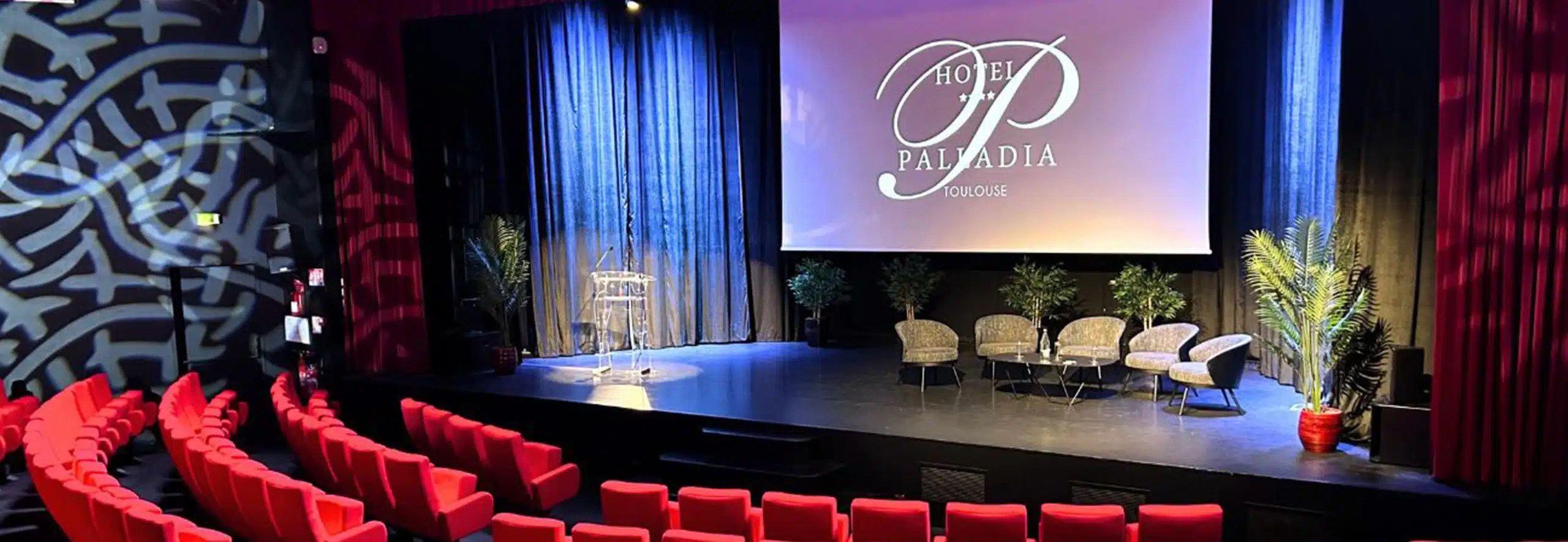 Une salle de conférence élégante avec des sièges rouges disposés en arc de cercle, face à une scène équipée d'un grand écran projetant le logo de l'Hôtel Palladia à Toulouse. L'ambiance est chaleureuse grâce à un éclairage doux en tons violets, créant une atmosphère professionnelle et accueillante.