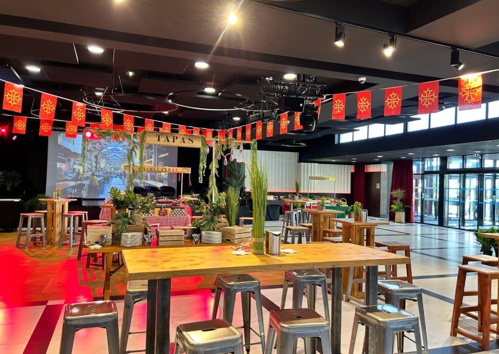 Décoration d'un espace événementiel de l'hôtel Palladia avec une ambiance chaleureuse et festive inspirée du sud de la France. Espace décoré avec des tables en bois, des chaises hautes en métal, un bar à tapas et des guirlandes de drapeaux aux couleurs occitanes. Parfait pour les réceptions, séminaires et soirées privées.